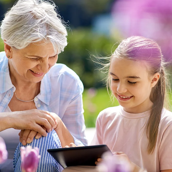 Eine ältere Dame mit grauen kurzen Haaren und ein junges Mädchen mit Zopf in der Natur, beide schauen auf ein Tablet.