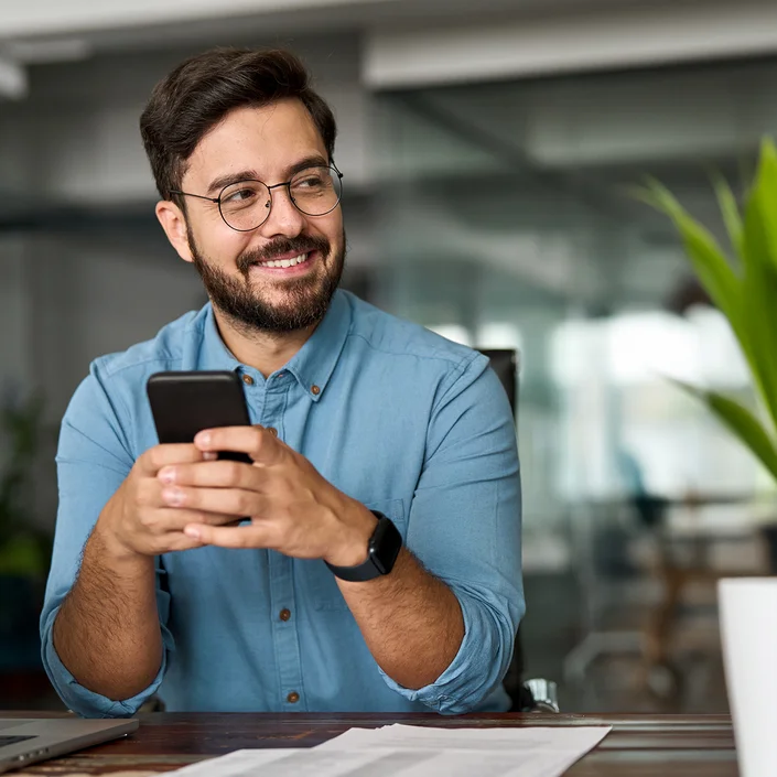 Ein Mann mit Brille in einem blauen Hemd sitzend an einem Tisch, er hält ein Smartphone in der Hand.