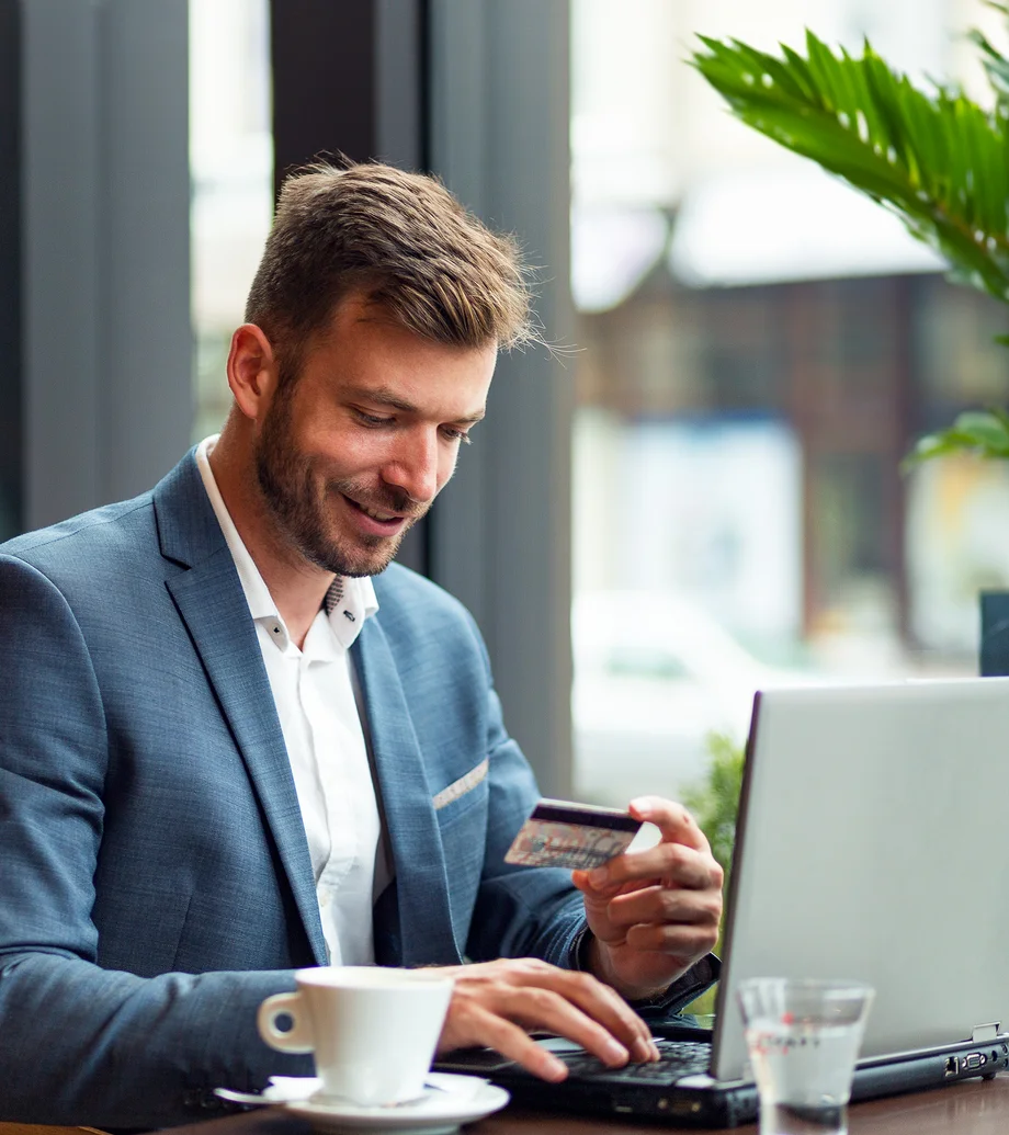 Ein Mann im Business-Outfit sitzend an einem Tisch: in der Hand hält er eine Bezahlkarte, am Tisch vor ihm steht ein Laptop, ein Kaffee und ein Glas Wasser.