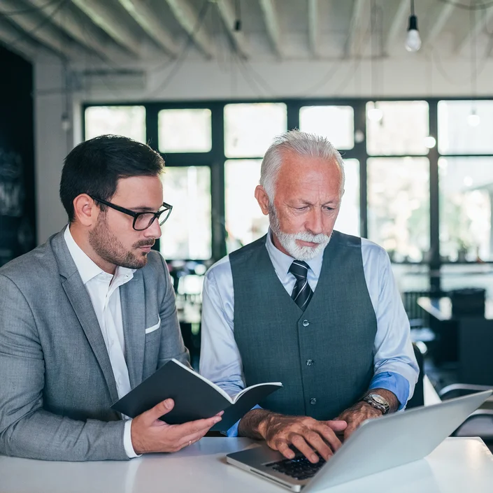 Foto von zwei Personen: Ein älterer und ein jüngerer Mann im Business-Outfit - beide schauen auf den Bildschirm eines Laptops. Der jüngere Mann hält ein Notizbuch in der Hand.