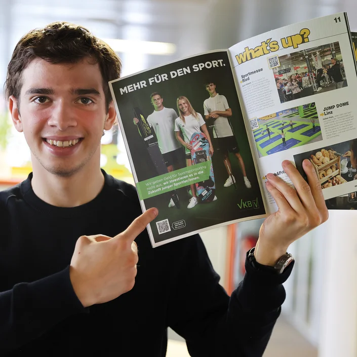 Ein Mann im schwarzen Longsleeve-Shirt, der das Magazin der 4youCard in die Höhe hält und mit dem Finger auf ein Inserat der VKB zeigt.
