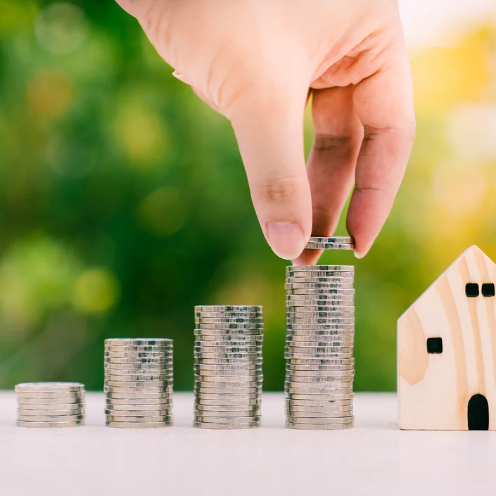 Foto mit 4 unterschiedlich großen Münzstapeln und einer Hand, die gerade Münzen auf dem höchsten Stapel ablegt, daneben steht ein kleines Holzhaus.
