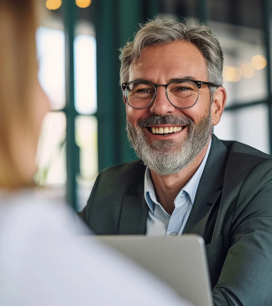 Foto eines Herren mittleren Alters mit Dreitagebart und Brille, er lächelt und sitzt vor einem Laptop - gegenüber eine Dame, die man nur verschwommen sieht.