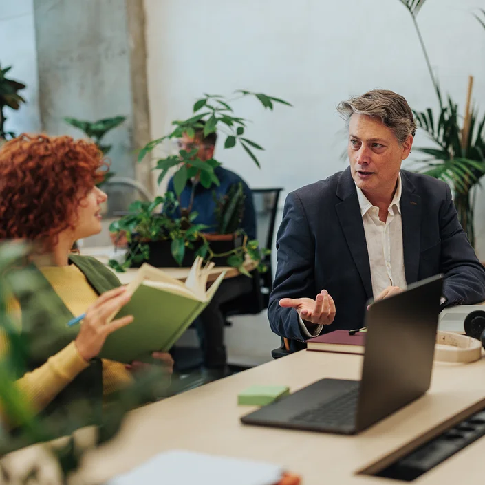 Foto einer Besprechungssituation in einem Großraumbüro: Ein Mann und eine Frau im Business-Outfit, die Dame hält einen Stift und ein Notizbuch in der Hand. Der Herr erklärt gerade etwas. Im Hintergrund sieht man weitere Kollegen und Pflanzen.