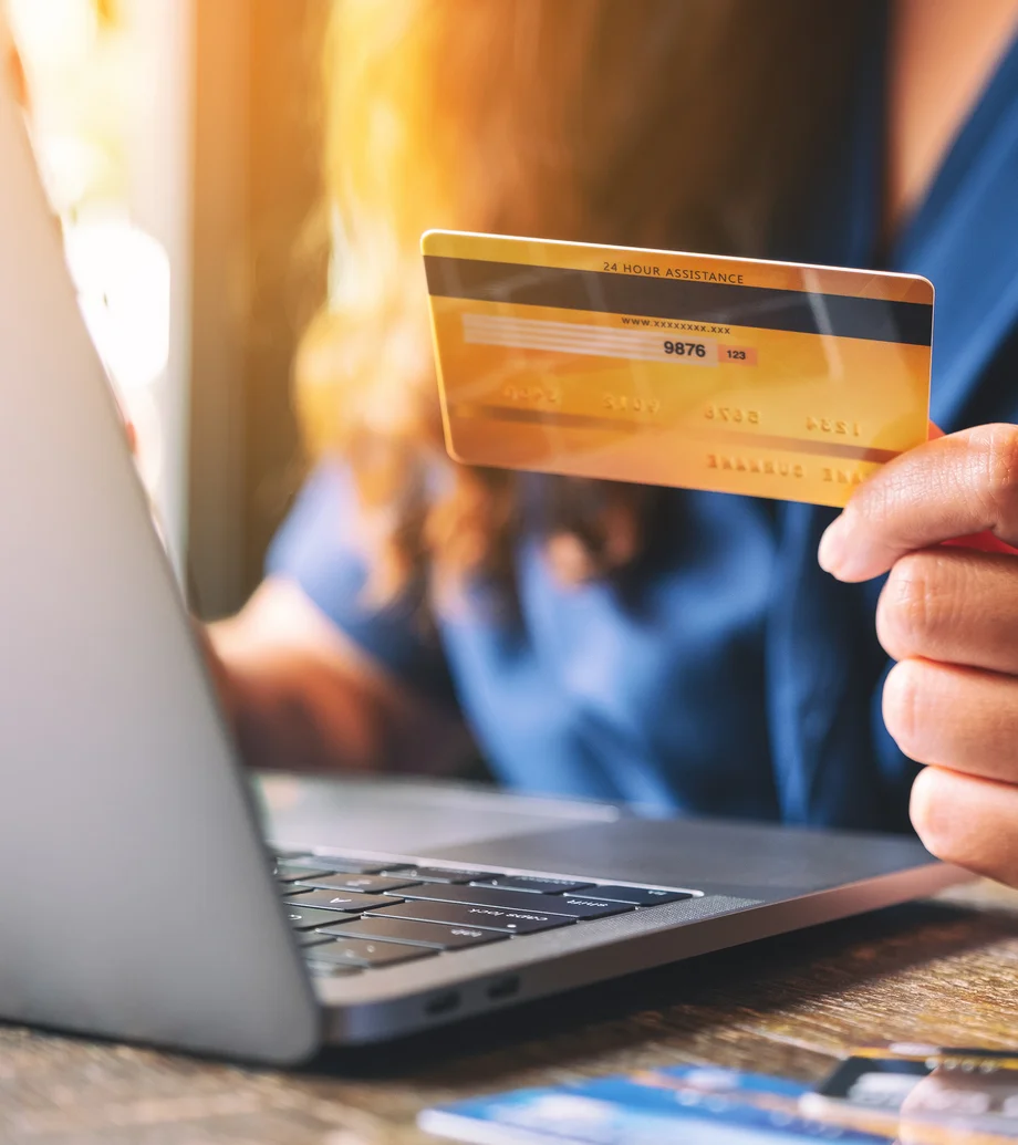 Close-up: Eine Frau mit blauem Shirt hält eine Bezahlkarte in ihrer Hand, sie sitzt vor einem Laptop.