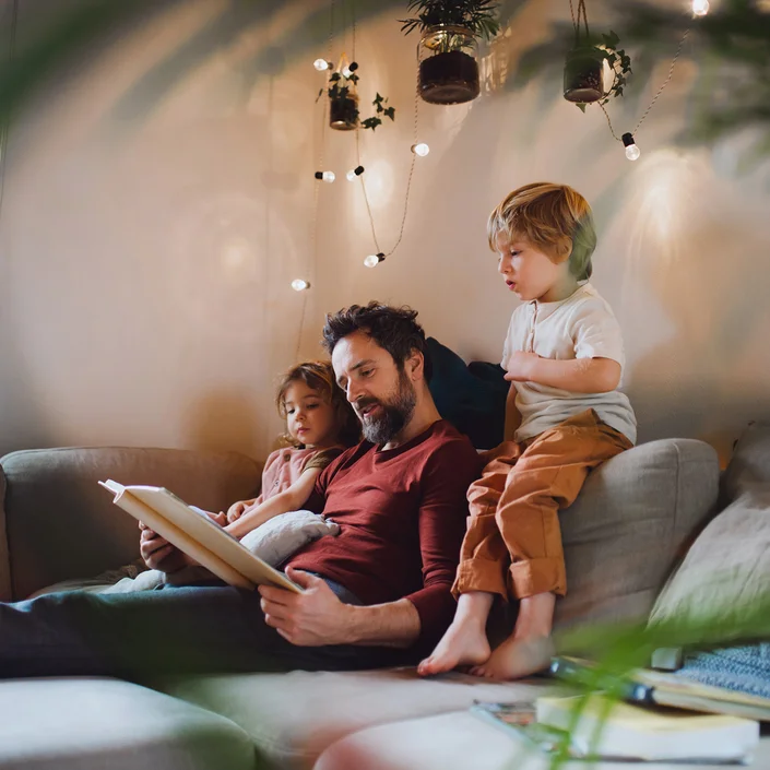 Foto eines Vaters mit seinen zwei kleinen Kindern auf der Couch, er liest den Kindern gerade etwas vor.