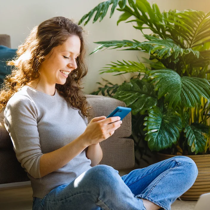 Foto einer Frau, die vor der Couch am Boden sitzt - sie hat ein Smartphone in der Hand. Im Hintergrund sieht man eine Pflanze.