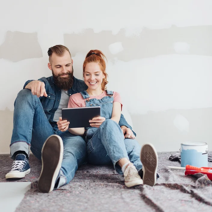 Foto eines Pärchen, das am Boden in einer Wohnung sitzt - die Frau hat einen Zopf und hält ein Tablet in der Hand. Neben ihnen steht ein Farbeimer und ein Pinsel.