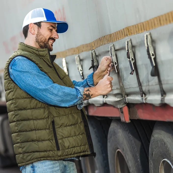 Ein Mann steht neben einem großen Lkw mit Plane und sichert die seitlichen Spanngurte. Er trägt eine blaue Jeans, eine olivgrüne Steppweste über einem Jeanshemd sowie eine blau-weiße Trucker-Cap.
