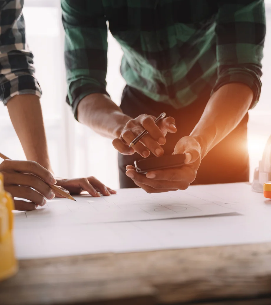 Foto von Händen, die einen Stift und ein Smartphone halten, im unteren Bereich des Bildes sieht man Bauunterlagen, die auf einem Tisch ausgebreitet sind.