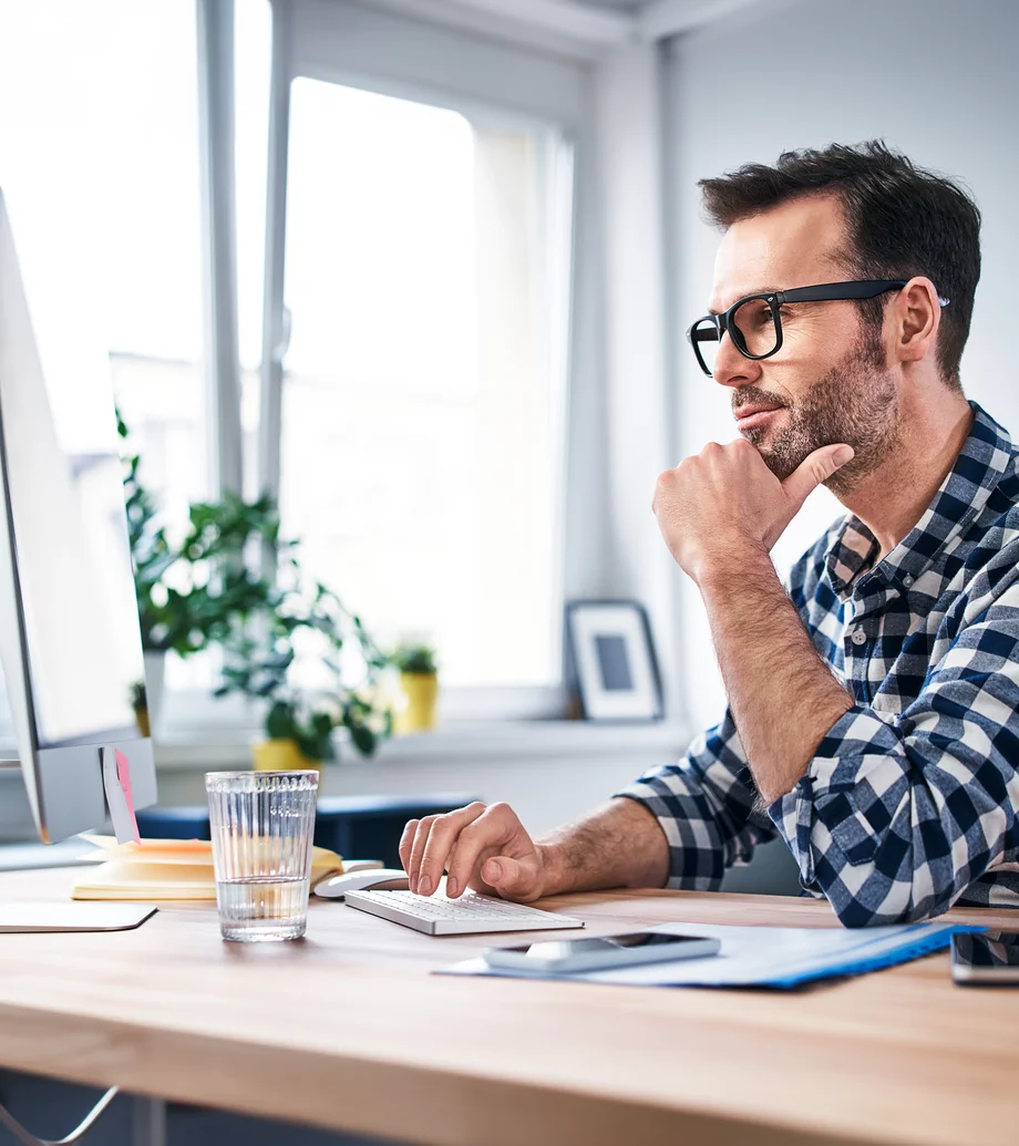 Ein Mann mit kariertem Hemd und Brille am Schreibtisch vor seinem PC.