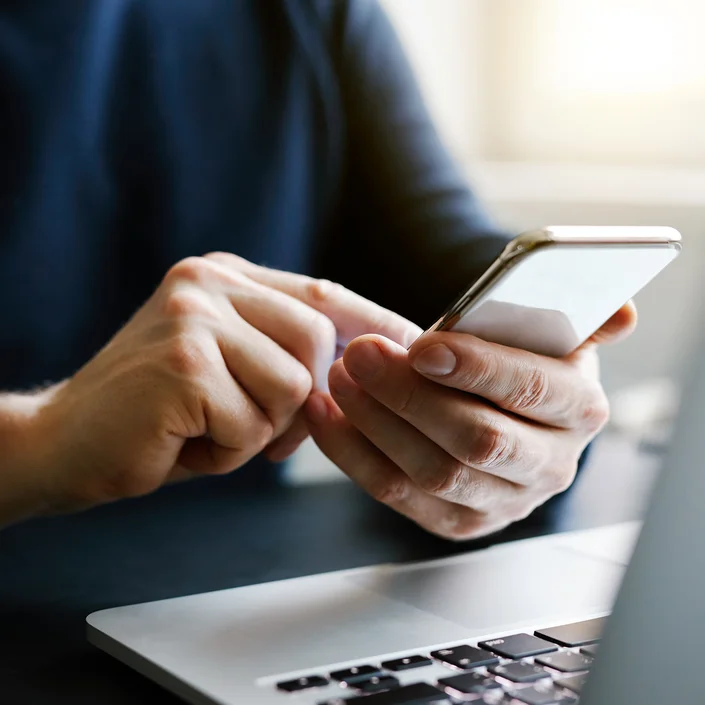 Close-up-Foto: Ein Mann sitzt vor einem Laptop - zu sehen sind seine Hände, die ein Smartphone halten.