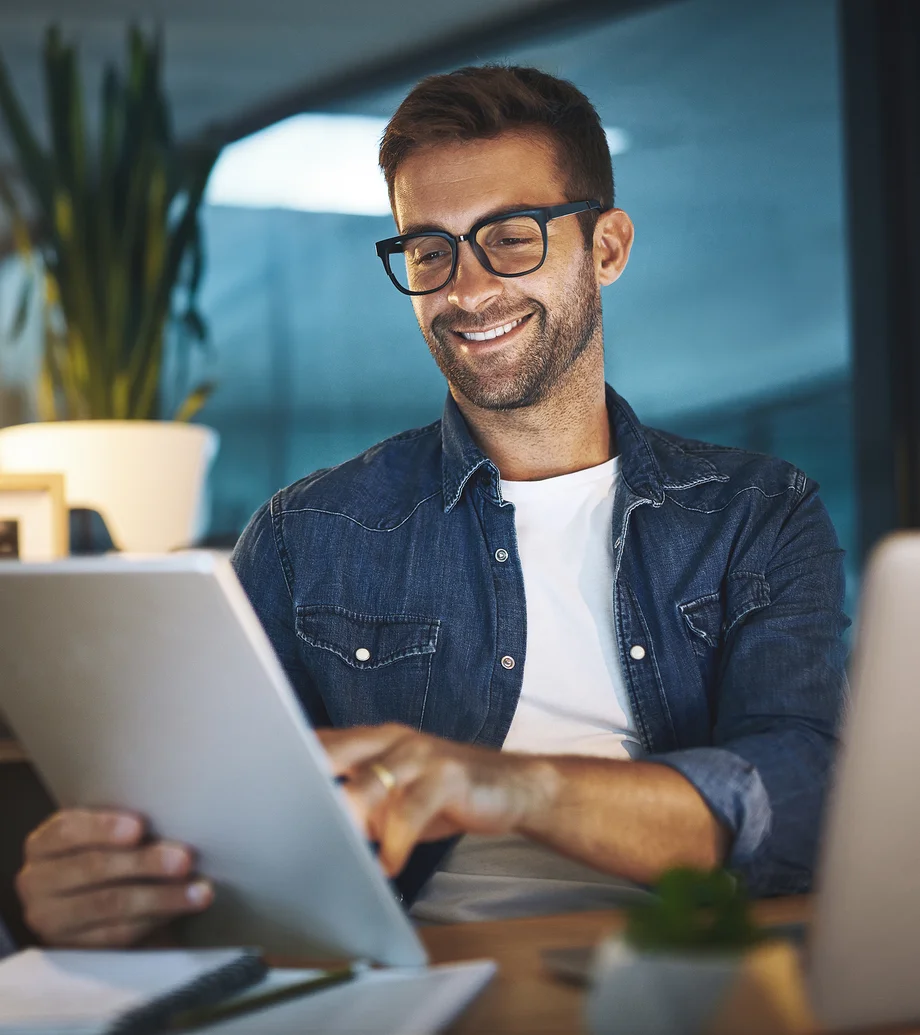 Ein Mann mit Brille und Jeanshemd sitzt mit einem Tablet an einem Tisch, auf dem Unterlagen liegen.