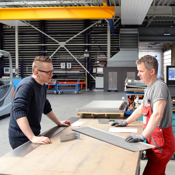Foto aus einer Werkstatt, 2 Handwerken stehen bei einer Werkbank zusammen und beraten sich.