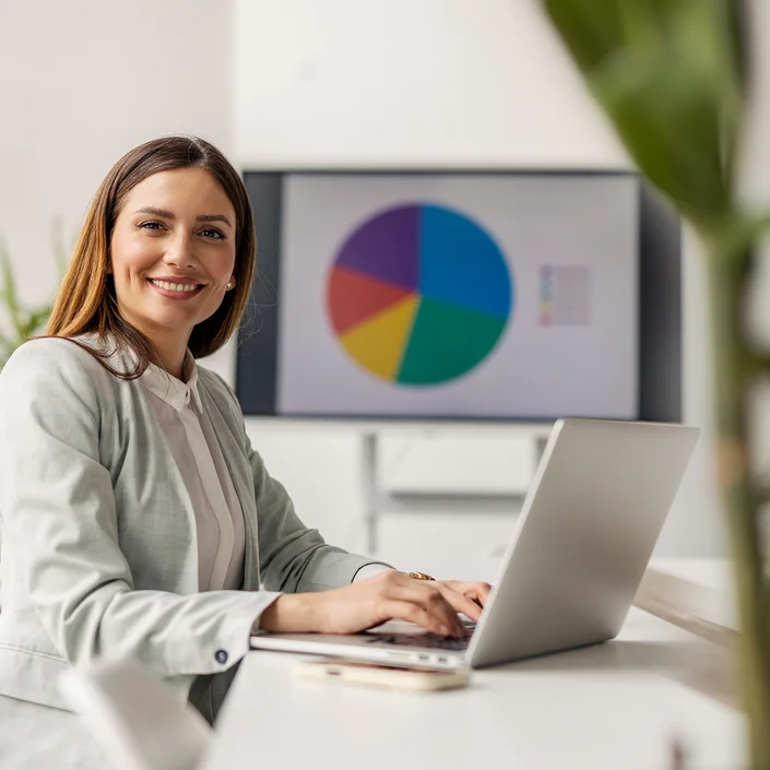 Foto einer Frau mit braunen langen Haaren im Business-Outfit, an einem Tisch sitzend, mit Laptop. Im Hintergrund ist ein Tortendiagramm auf einem Bildschirm zu sehen.