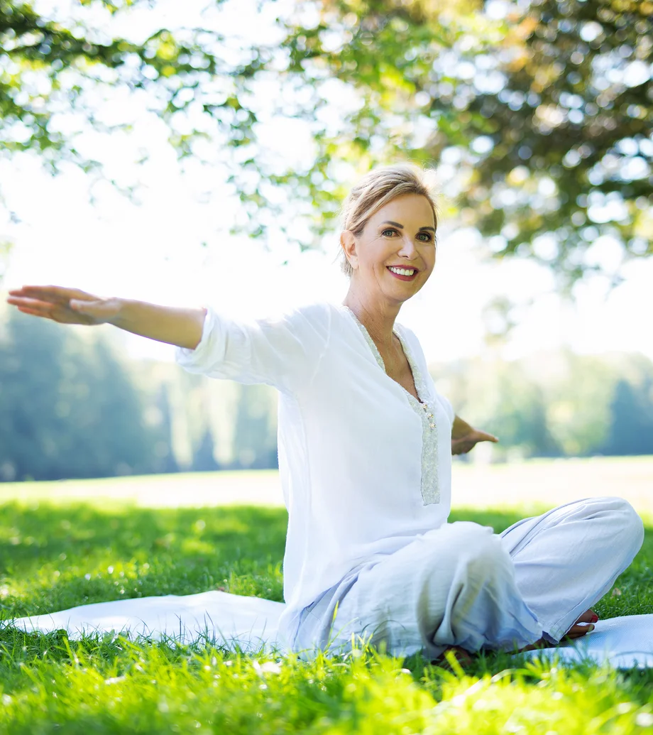 Eine Frau mit weißem Gewand auf einem Handtuch in der Natur beim Yoga.