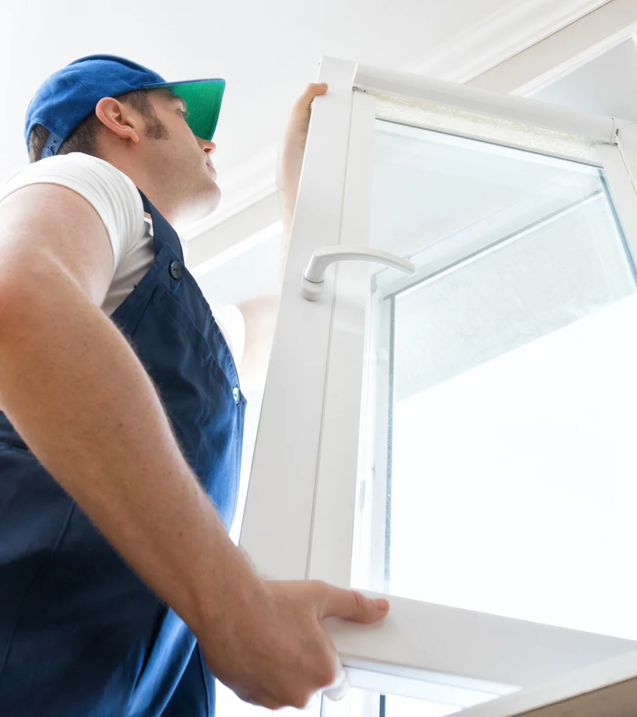 Foto eines Handwerkers im Blaumann und mit Kappe, der gerade ein Fenster einhängt.