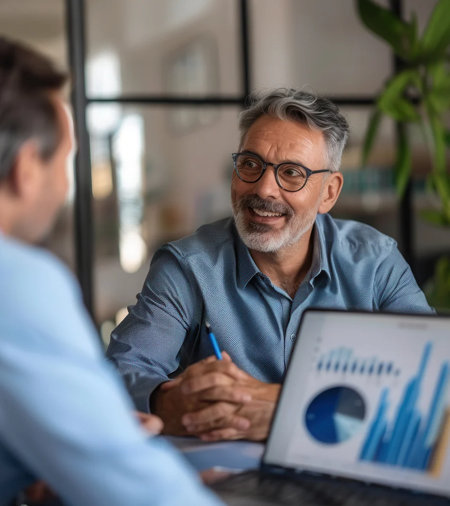 Foto eines Beratungsgespräch: 2 Männer sitzen an einem Tisch, der eine hat einen Laptop vor sich und schaut sich Auswertungen am Bildschirm an.