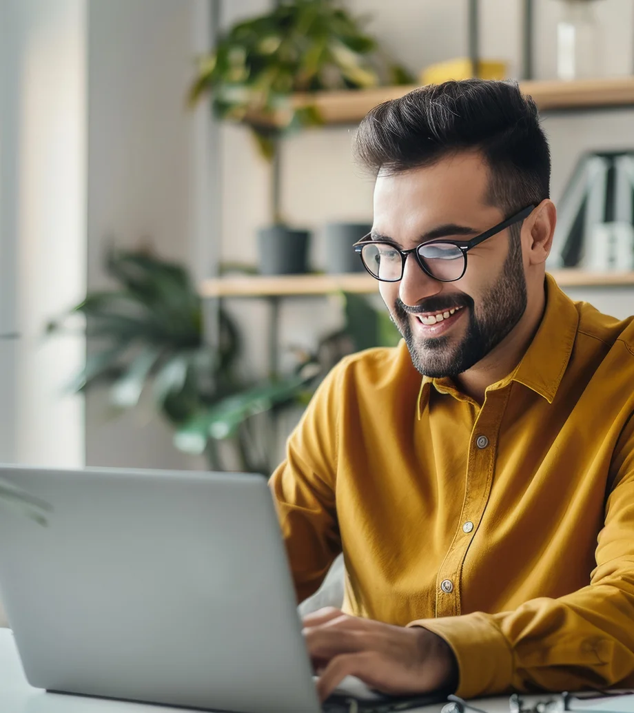 Ein Mann mit Brille und orangefarbenem Hemd sitzt am Tisch vor einem Laptop, im Hintergrund ist ein Regal zu sehen, auf dem Pflanzen stehen.